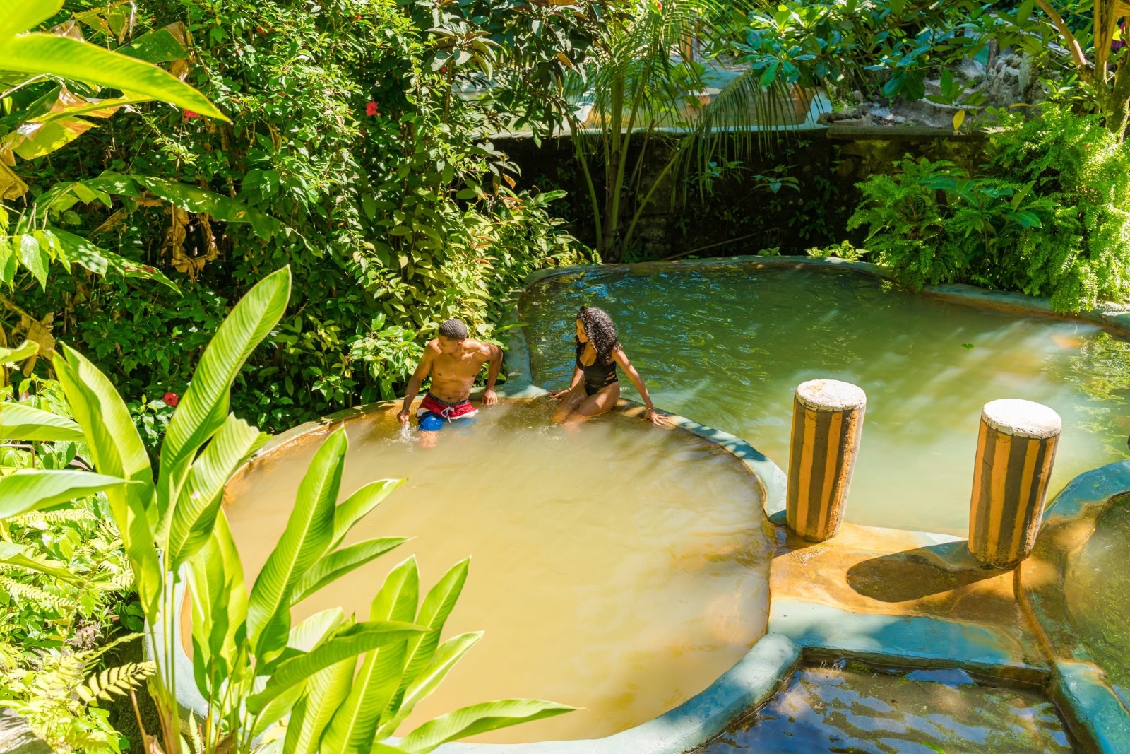 Two travellers wading in tiered volcanic mineral pools ringed by tropical plants